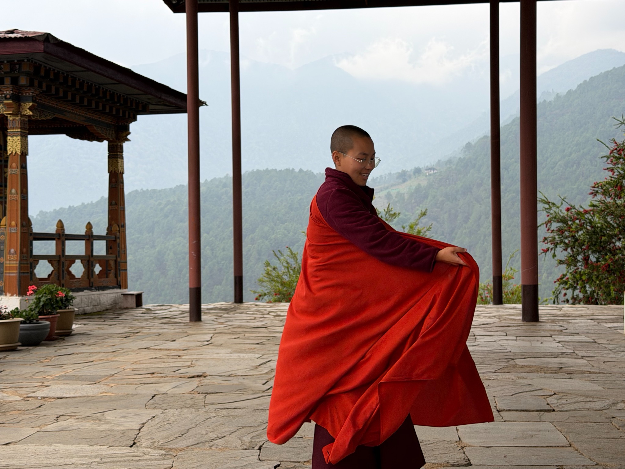 Monastic figure in a red robe standing in a Bhutanese pavilion against mountains