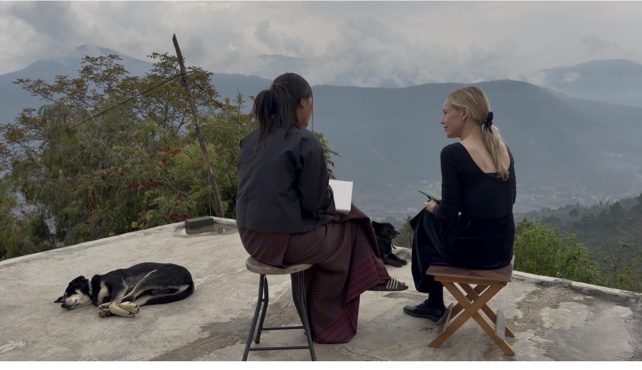 Two women seated in conversation overlooking a Bhutanese valley with dogs nearby