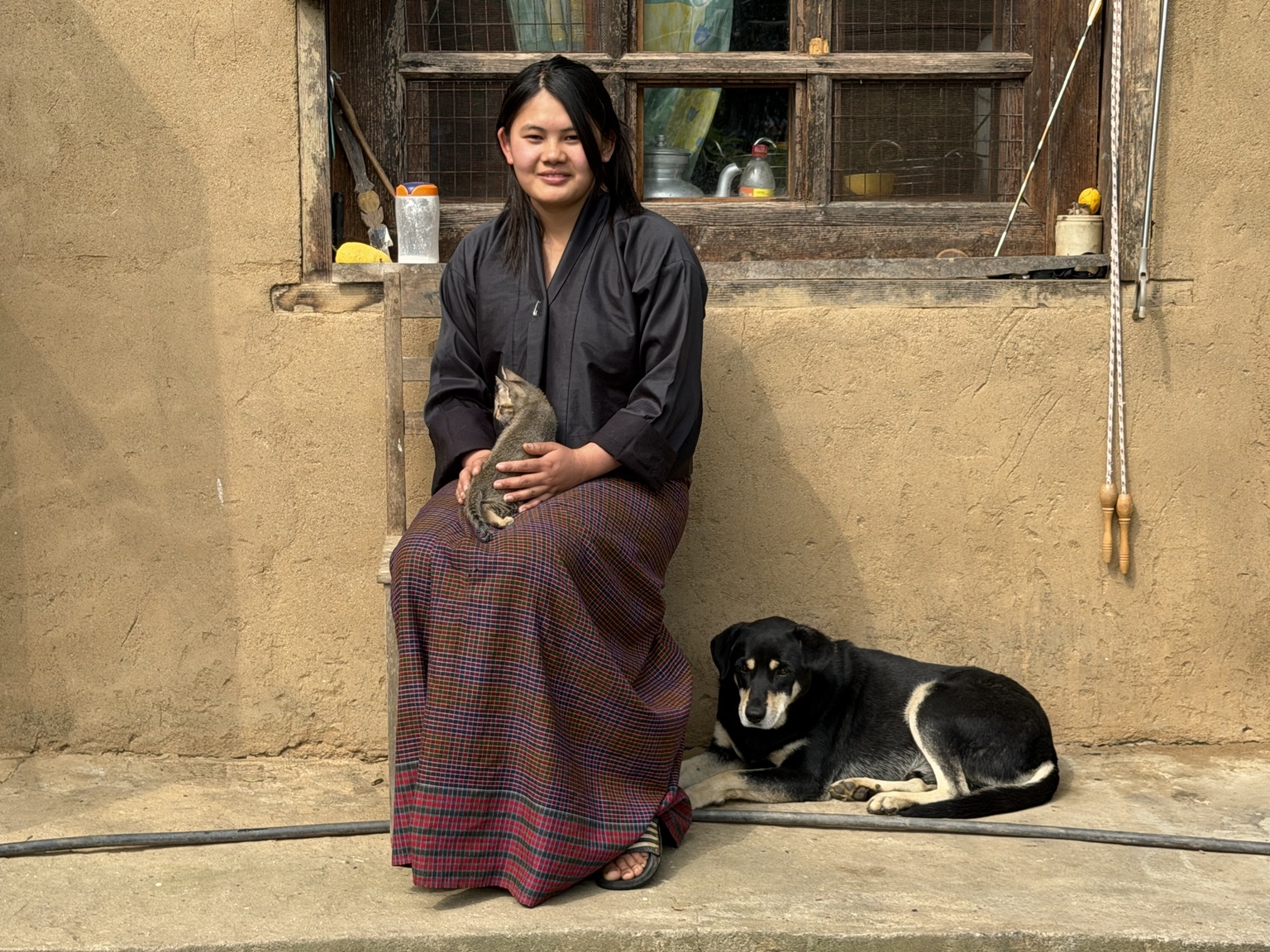 Bhutanese woman seated outside a traditional house holding a cat, with a dog beside her